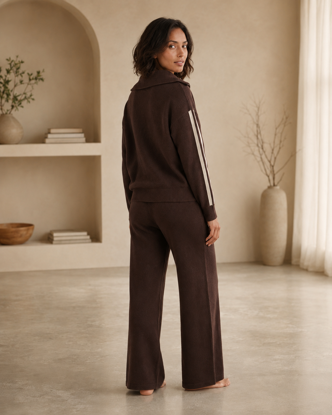 A woman stands barefoot in a warm neutral room, wearing a dark brown matching lounge set with a zip-front hoodie and wide-leg pants. The hoodie has white vertical stripes down the sleeves.