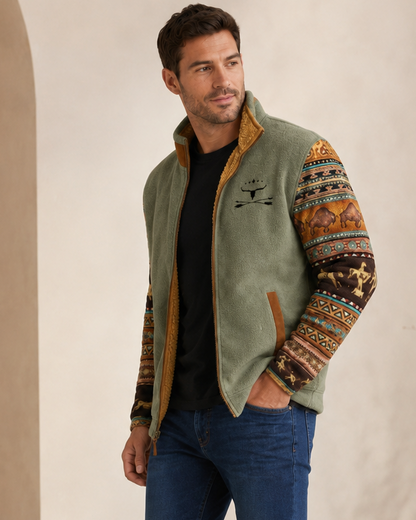 A man wearing a sage green fleece vest with patterned sleeves, blue jeans, and brown leather boots, standing in a warm, neutral-toned interior setting.