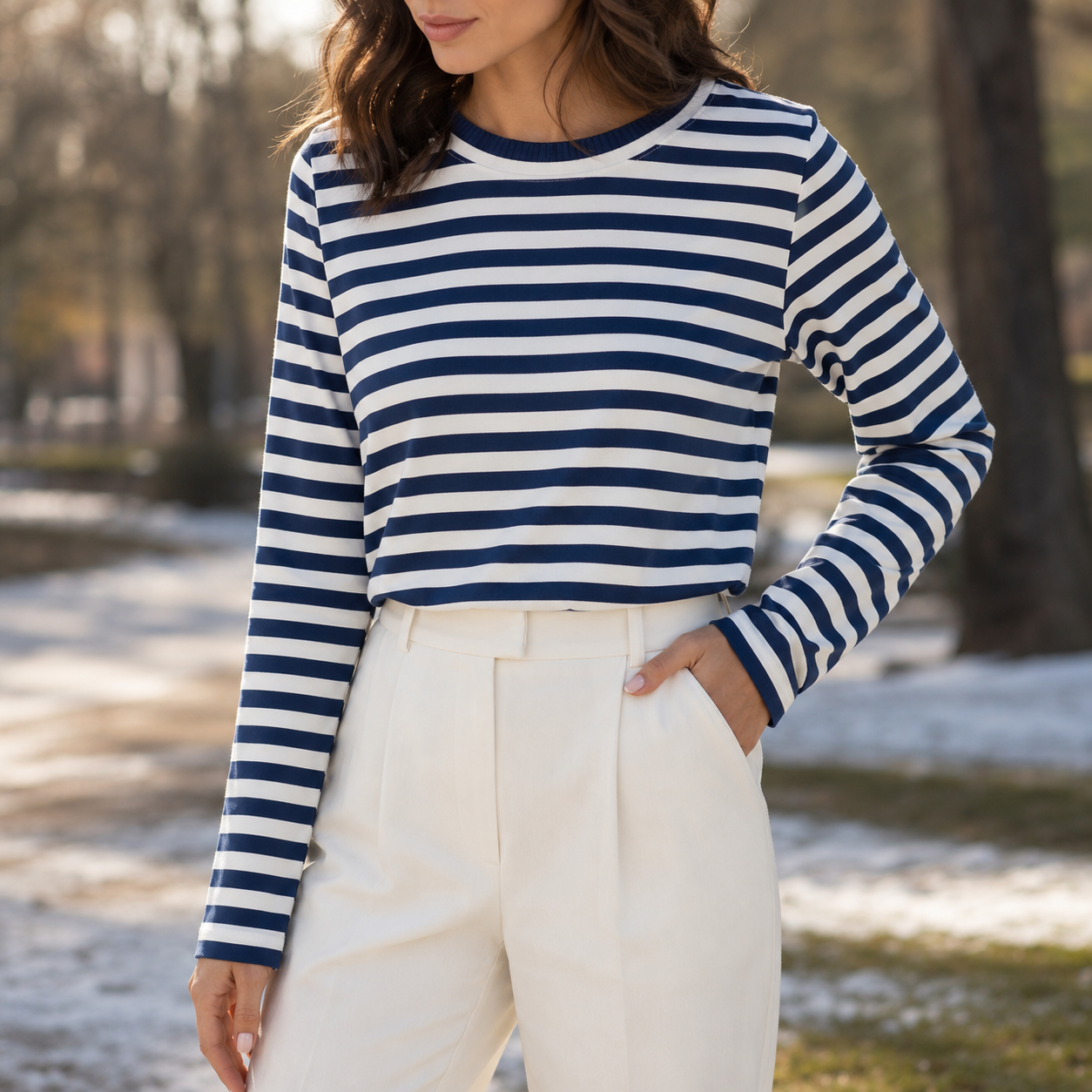 A woman wearing a navy and white horizontally striped long-sleeve top with white trousers, standing outdoors in a park-like setting with blurred trees and ground patches of snow.
