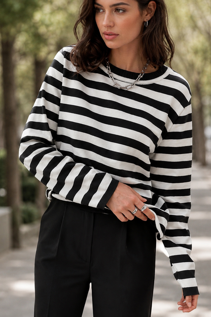 A woman wearing a black and white horizontally striped long-sleeve knit top and black trousers, standing on a tree-lined sidewalk.